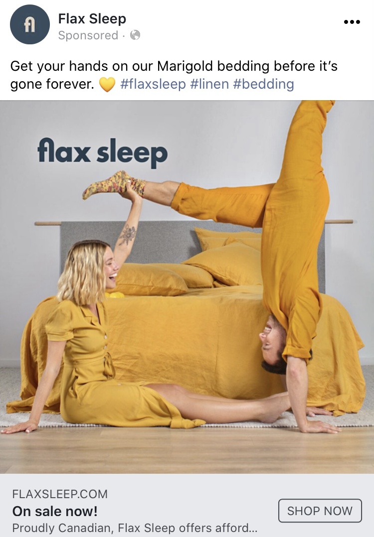 Two people doing acro yoga dressed in yellow clothing in front of a bed with the same colour sheets.